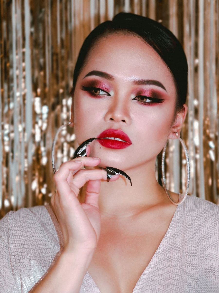 East Asian woman with red lipstick, bold eye makeup and hoop earrings against a silver tinsel backdrop