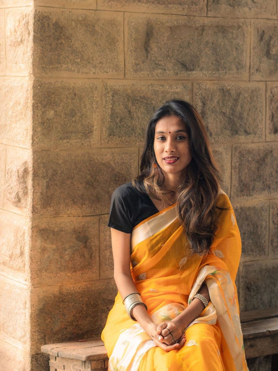 Indian woman in a yellow silk saree seated on a bench beside a stone wall