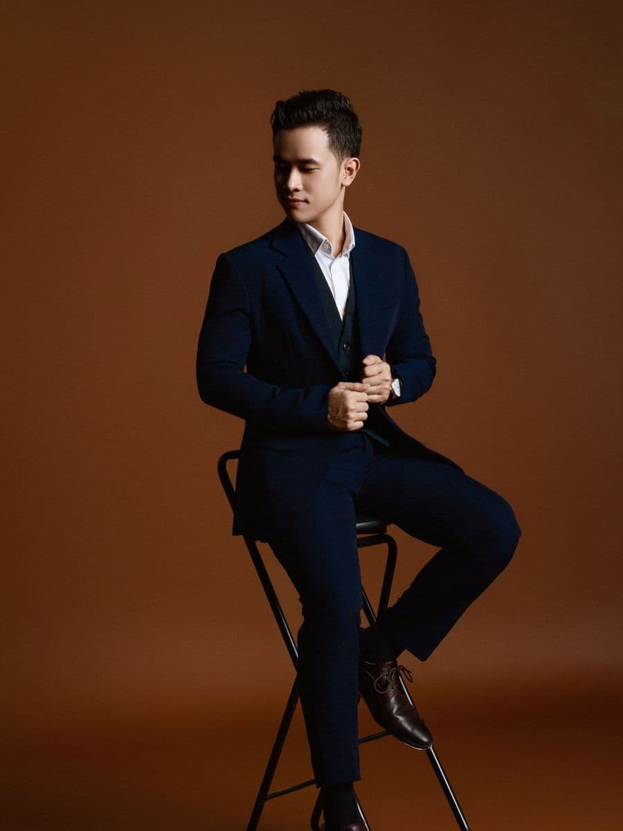 Man in a navy suit seated on a tall stool against a brown studio backdrop