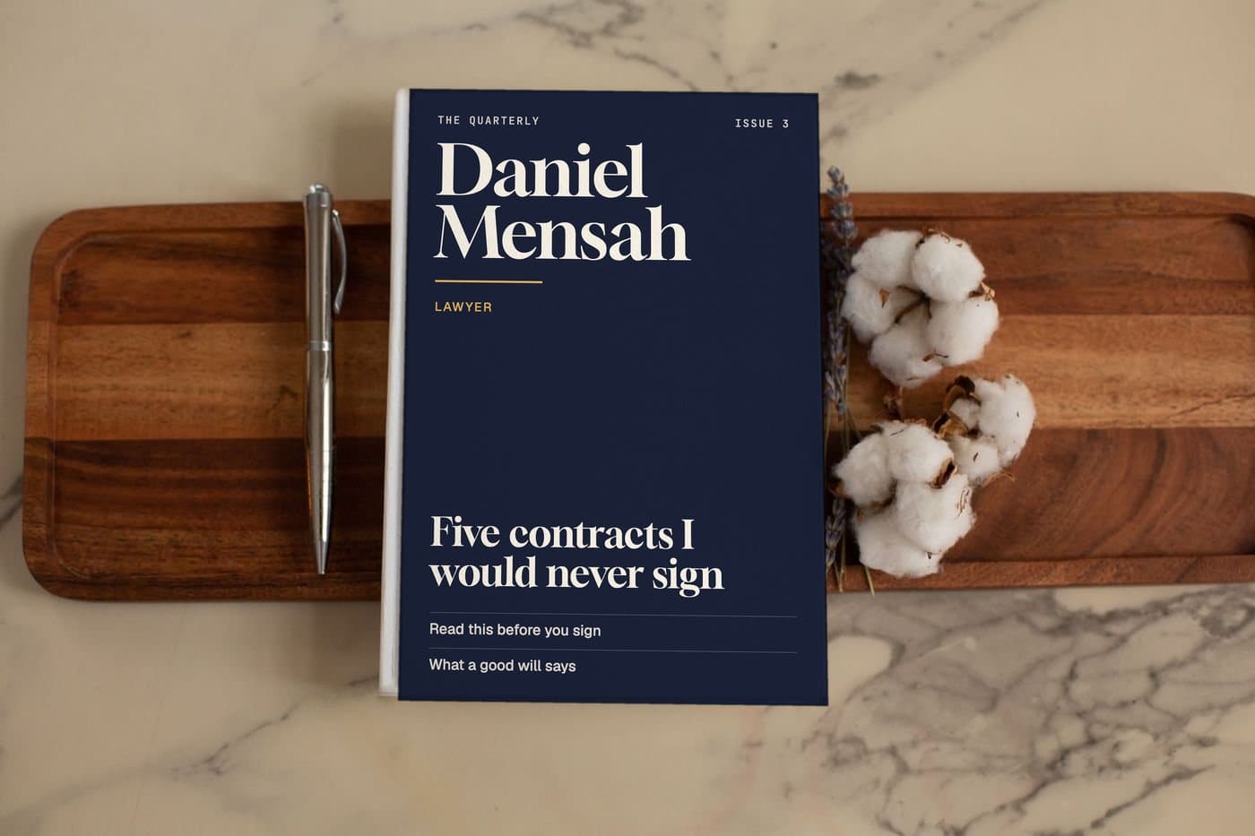 A sample quarterly magazine with Daniel Mensah on the cover. Setting: a blank white book on a marble table with a wooden tray, a pen and cotton flowers.