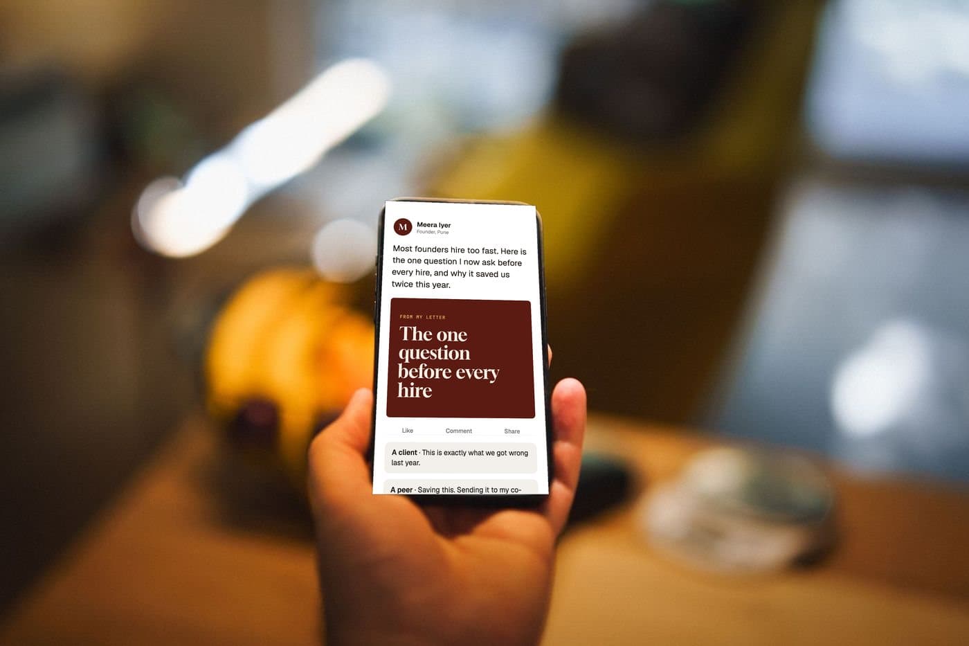 A phone showing a sample post by Meera Iyer. Setting: a hand holding a smartphone with a blank screen above a café table in warm evening light.