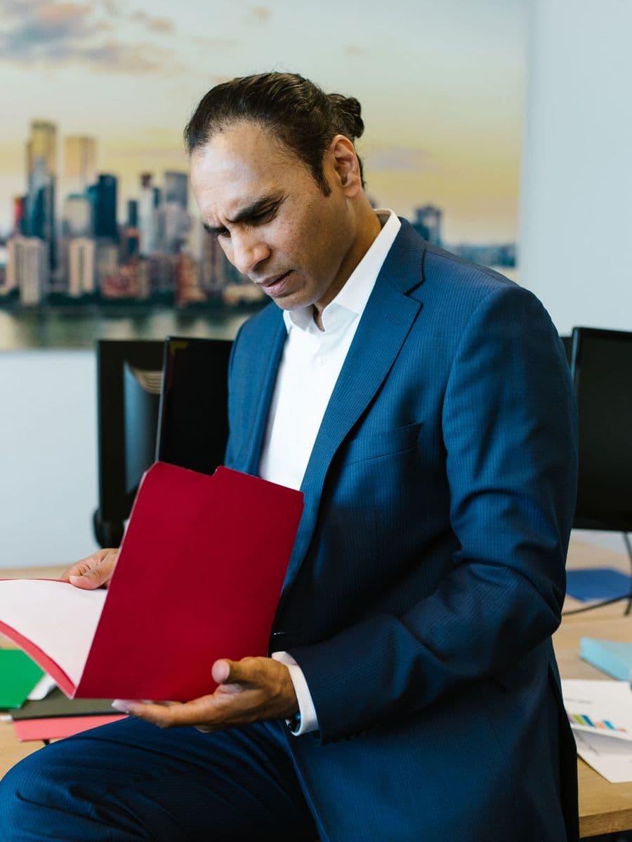 South Asian man in a blue suit reviewing a red file at an office desk