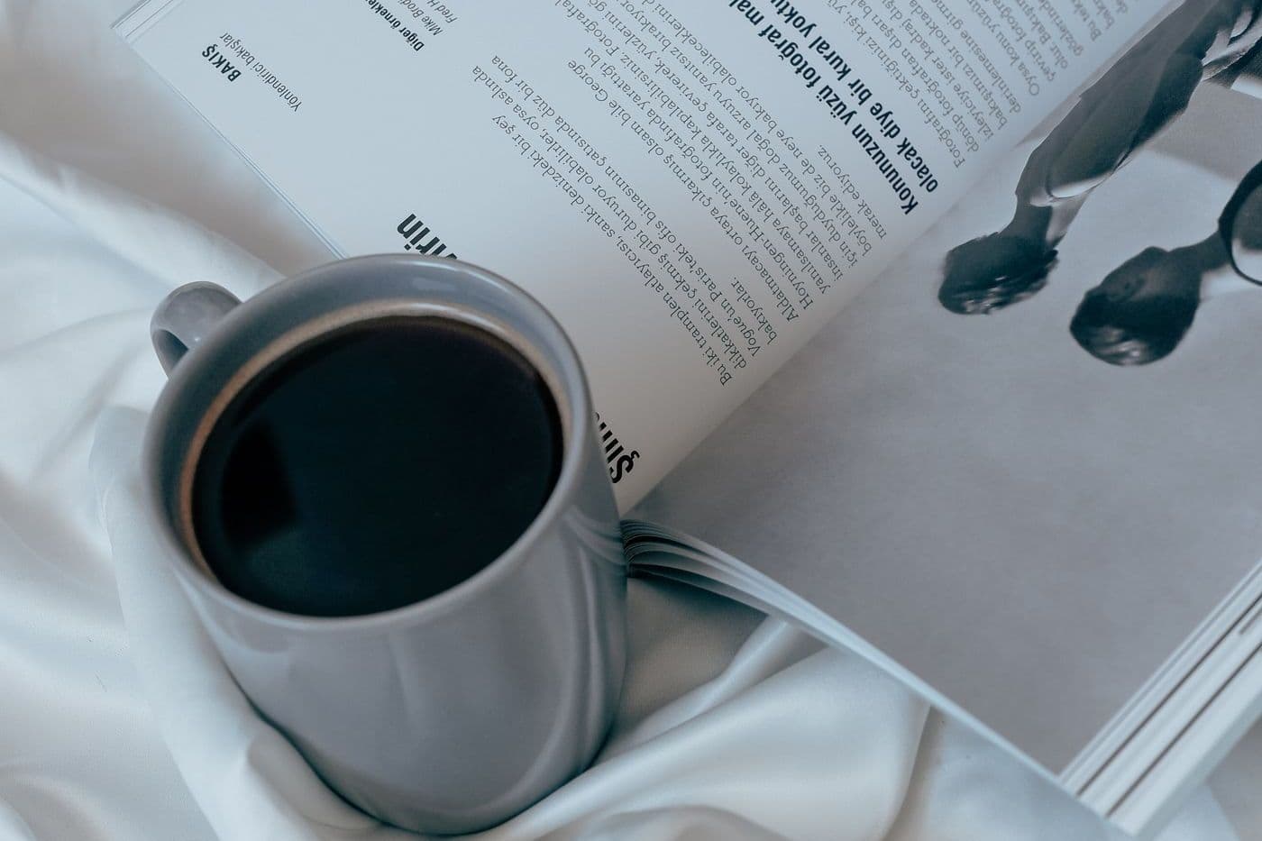 Open magazine and a mug of black coffee on white bed sheets